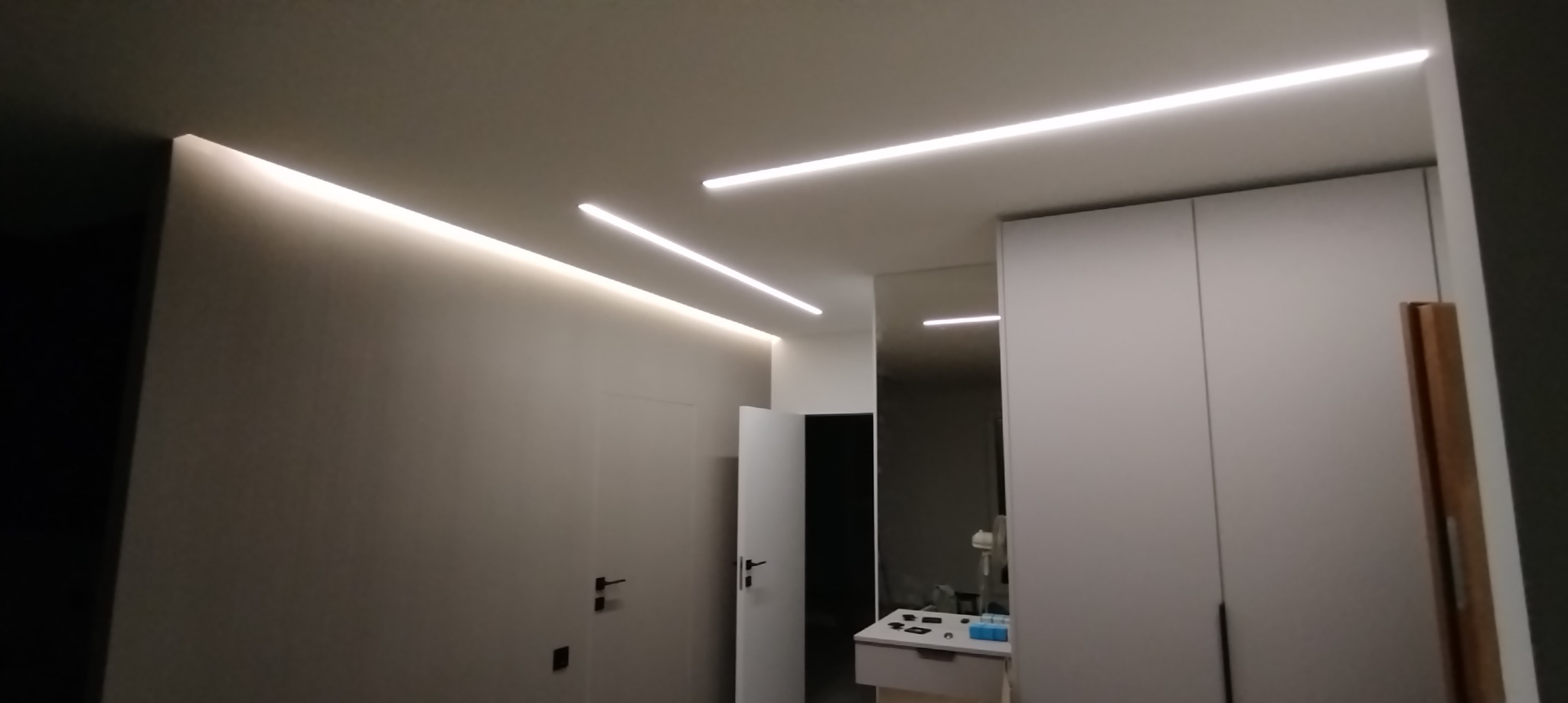 Nowoczesne oświetlenie LED w minimalistycznym wnętrzu. Białe ściany i sufit z subtelnymi liniami świetlnymi. Widoczne drzwi i fragment szafy w zabudowie.