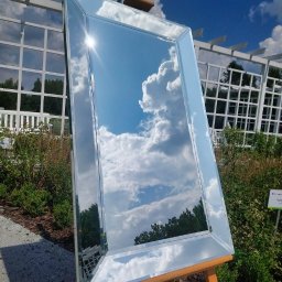 M-K Studio - Prostokątne lustro z fazowanymi krawędziami, ustawione na drewnianym stojaku malarskim, odbija błękitne niebo z białymi chmurami w ogrodowej scenerii.