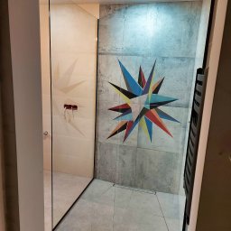 M-K Studio - Nowoczesna kabina prysznicowa walk-in z czarnymi profilami, wykończona płytkami z geometrycznym wzorem na ścianie. Minimalistyczny design, widoczny odpływ liniowy.