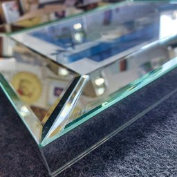 M-K Studio - Szklana tafla z fazowanymi krawędziami, leżąca na szarym materiale. Widoczne refleksy światła i otoczenia na powierzchni szkła. Detal wykończenia.