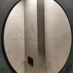 M-K Studio - Okrągłe lustro w czarnej ramie, odbijające wykończoną łazienkę z szarymi płytkami. Realizacja z Łasku.