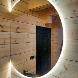 M-K Studio - Drewniana łazienka z okrągłym lustrem LED. Widoczna umywalka i bateria. Ściany i sufit wyłożone boazerią. W lustrze odbija się ciemne drzwi.