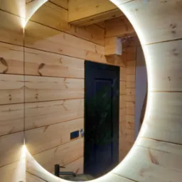 Drewniana łazienka z okrągłym lustrem LED. Widoczna umywalka i bateria. Ściany i sufit wyłożone boazerią. W lustrze odbija się ciemne drzwi.