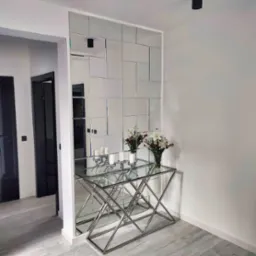 Ściana z lustrzanych płytek, tworząca geometryczny wzór. Na tle białych ścian, z konsolą ze stali i szkła, na której stoją świece i kwiaty.