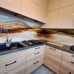 Nowoczesna kuchnia z drewnopodobnymi szafkami, ciemnym blatem i szklanym panelem dekoracyjnym w odcieniach brązu. Widoczny zlew, płyta gazowa i fragment podłogi.