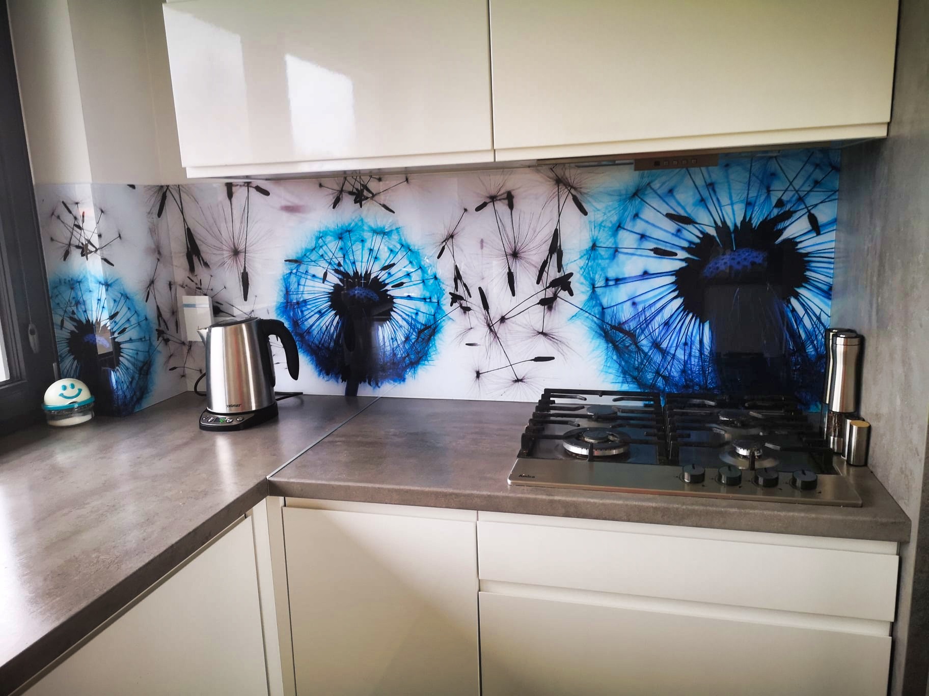 Szklany panel dekoracyjny z motywem dmuchawca w kuchni w Łasku. Nowoczesny design, białe szafki, szary blat i płyta gazowa. Estetyczne wykończenie.