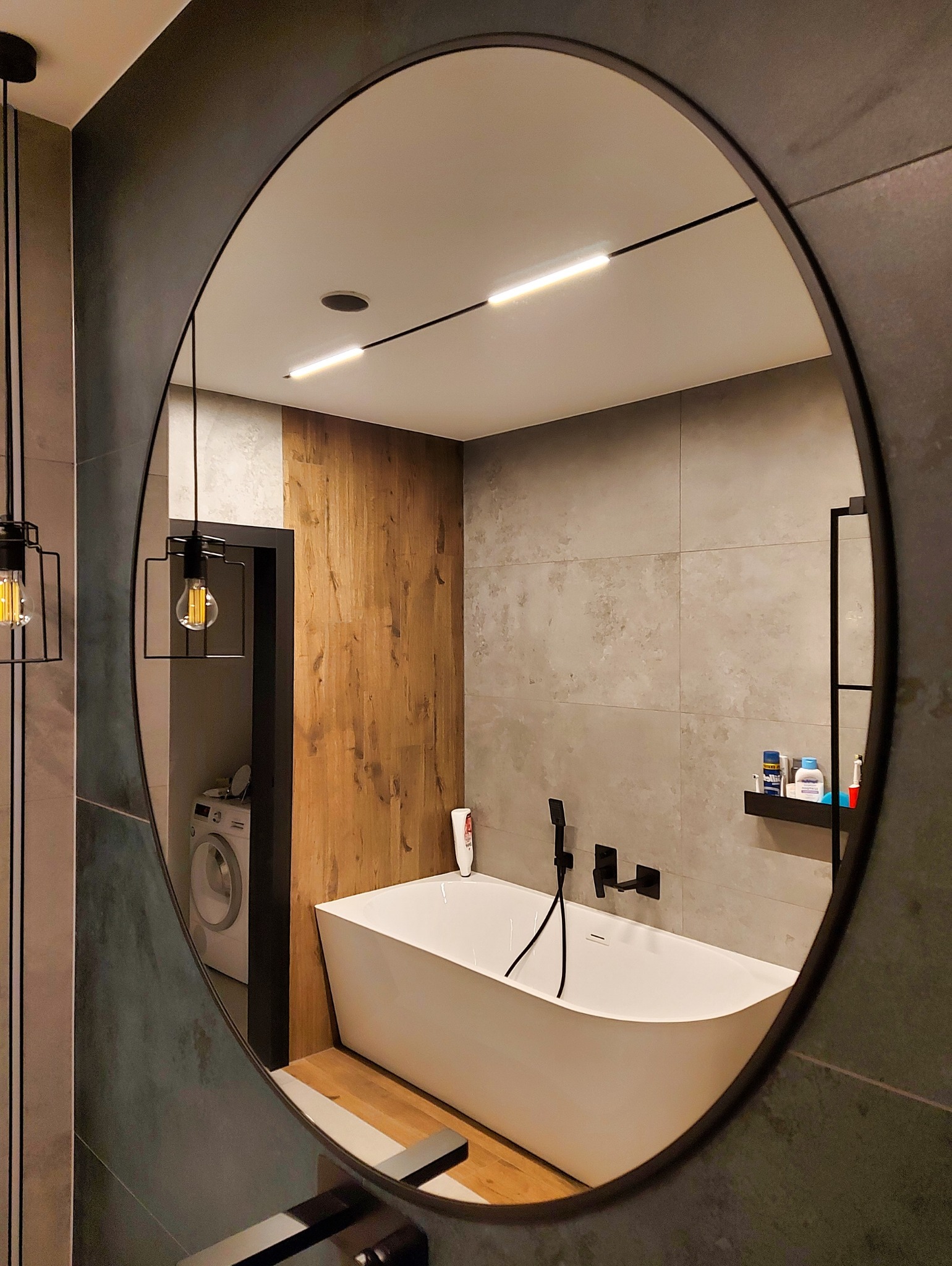 Nowoczesna łazienka odbita w okrągłym lustrze: biała wanna, szare płytki, drewniane akcenty, czarna armatura i designerskie oświetlenie. Widoczna pralka w tle.