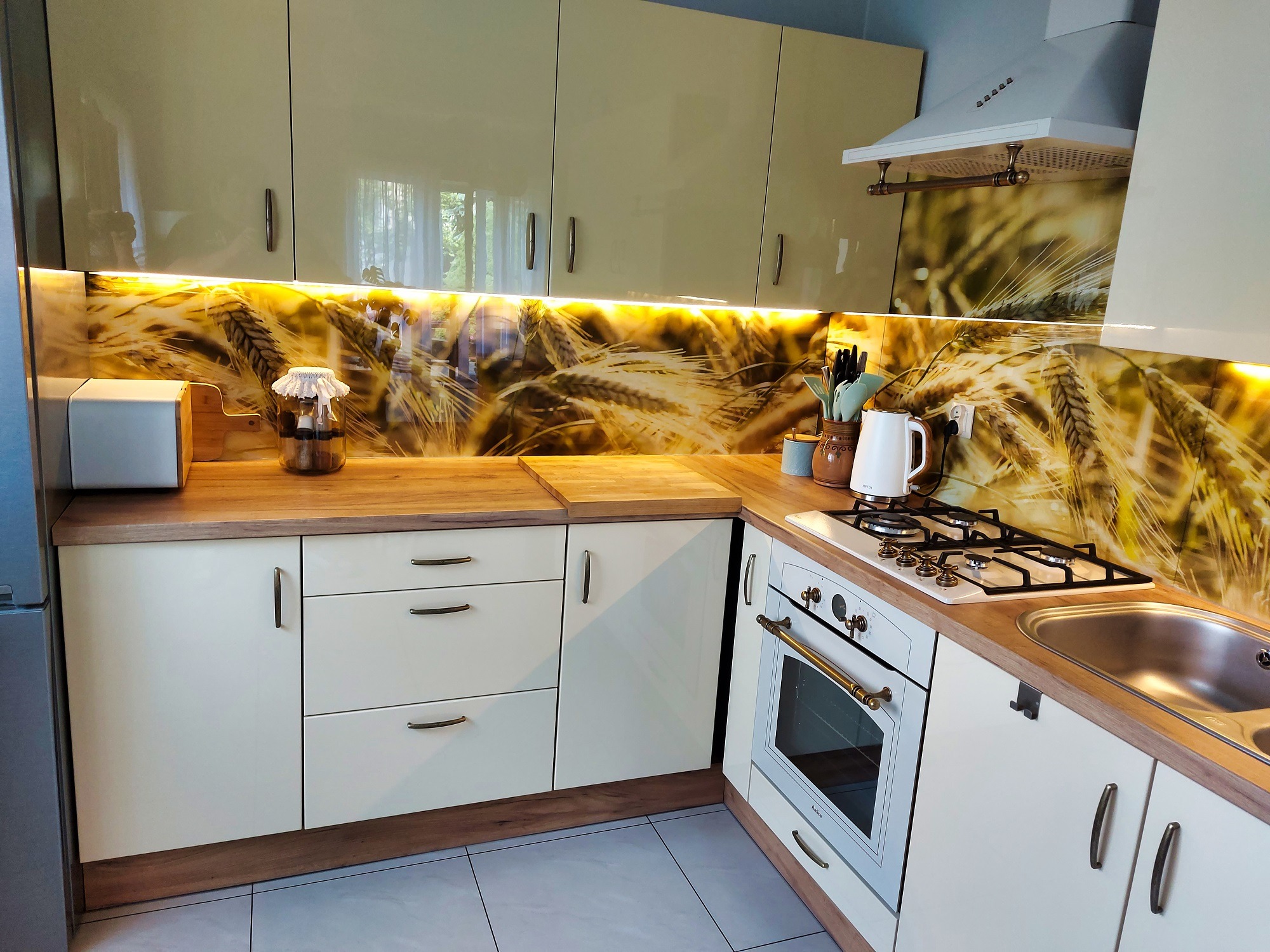 Jasna kuchnia z blatem imitującym drewno i panelem szklanym z motywem zboża. Biała zabudowa, piekarnik retro, okap. Oświetlenie LED podszafkowe.