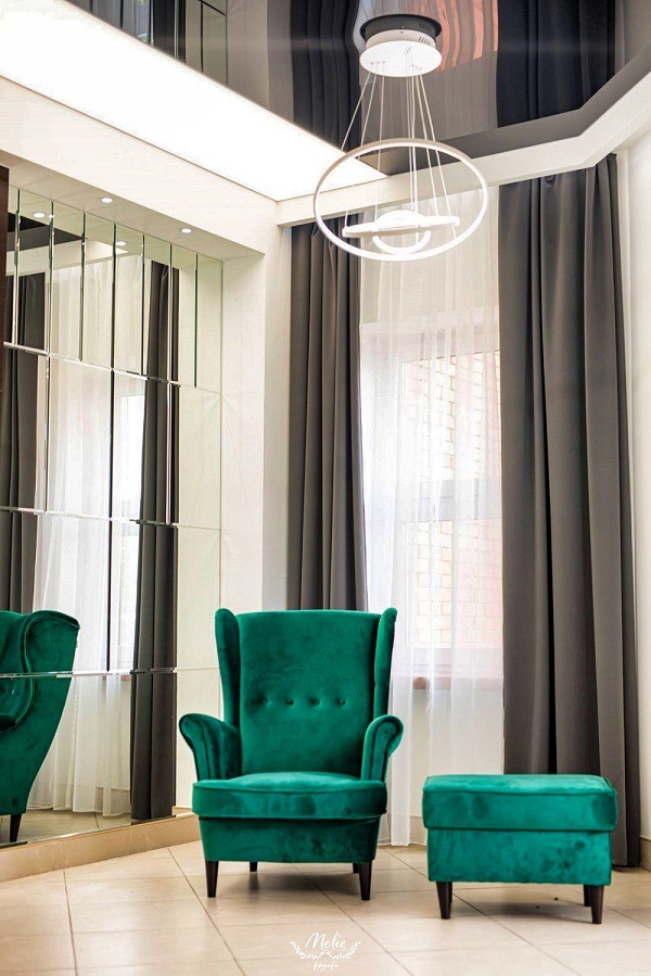 Szmaragdowy fotel typu uszak z podnóżkiem na tle ściany z lustrzanych paneli i zasłon. Nowoczesna lampa wisząca i błyszczący sufit.