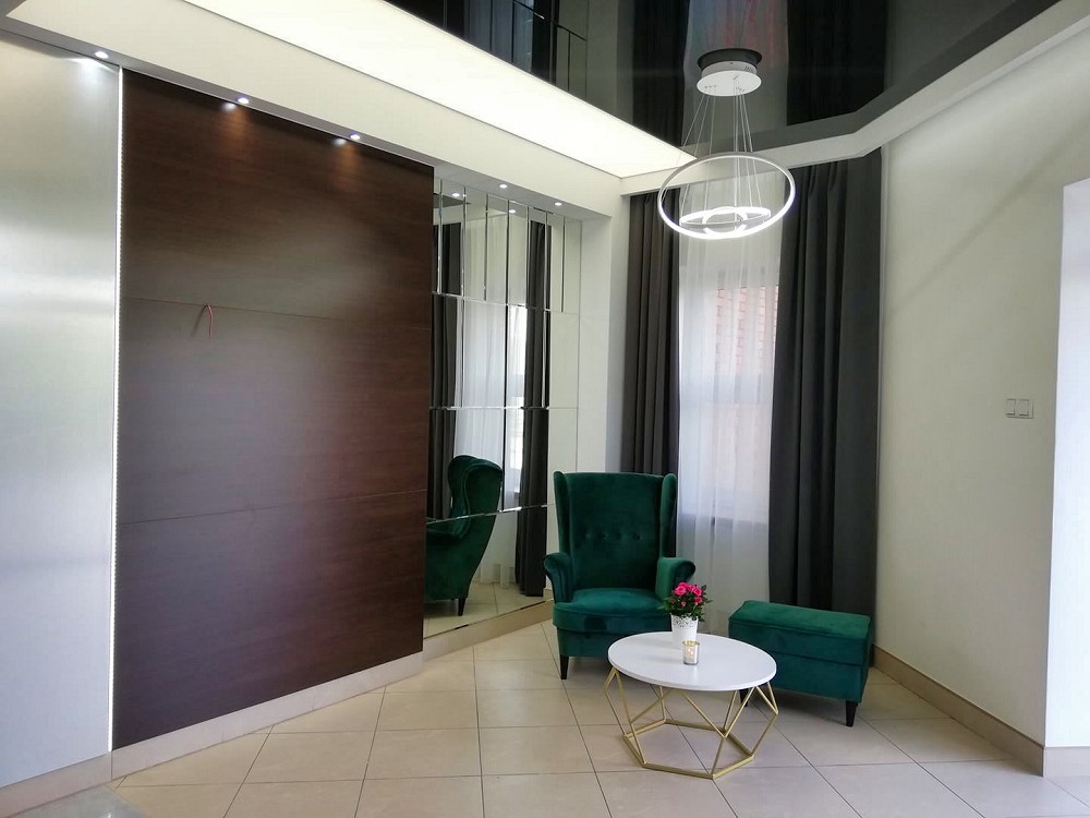 Elegancki salon z lustrami, drewnianą ścianą i zielonym fotelem. Nowoczesne oświetlenie sufitowe. Widoczne detale wykończenia i aranżacji wnętrza.