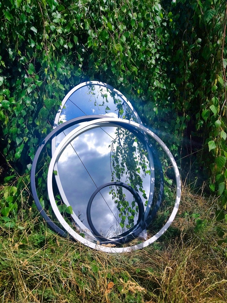 Okrągłe lustra w czarnych i białych ramach, ułożone na trawie, odbijają niebo. W tle zielone liście drzew, tworzące naturalną scenerię.