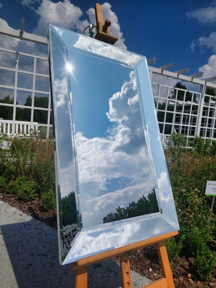 Prostokątne lustro z fazowanymi krawędziami, ustawione na drewnianym stojaku malarskim, odbija błękitne niebo z białymi chmurami w ogrodowej scenerii.