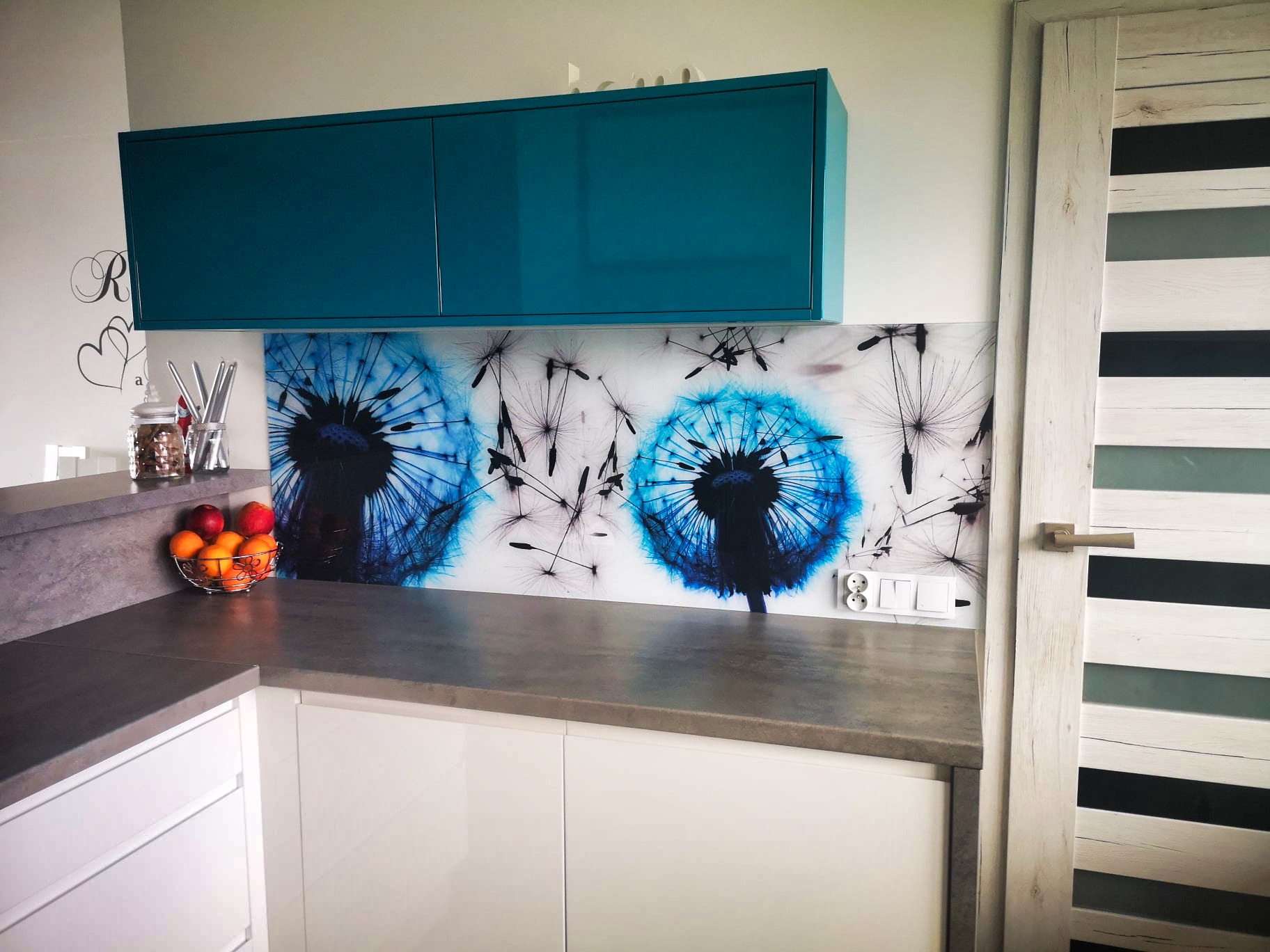 Biała kuchnia z turkusowymi szafkami i szklanym panelem z motywem dmuchawca. Blat w kolorze szarości. W tle drzwi z drewnianą ramą i poziomymi przeszkleniami.