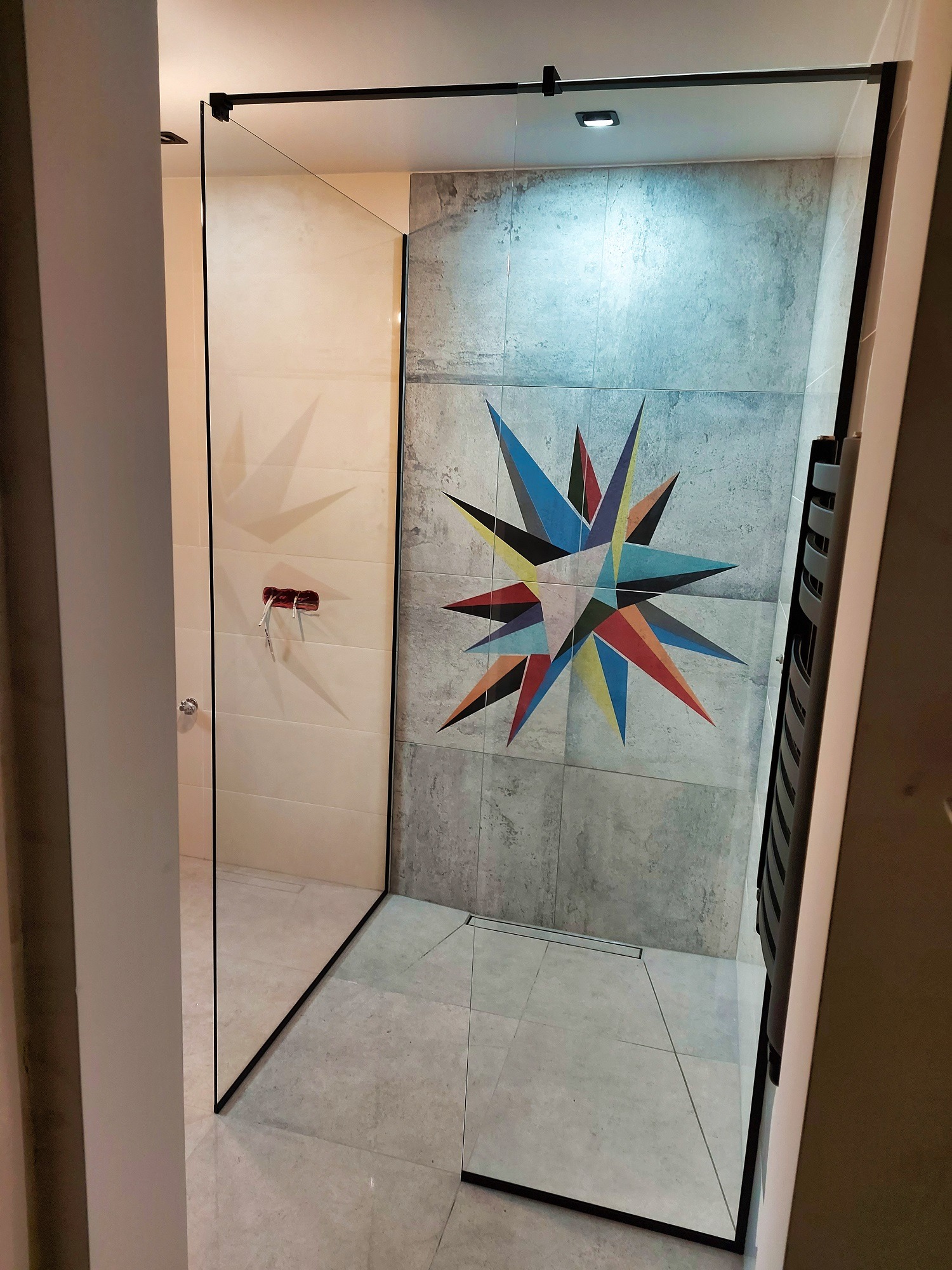 Nowoczesna kabina prysznicowa walk-in z czarnymi profilami, wykończona płytkami z geometrycznym wzorem na ścianie. Minimalistyczny design, widoczny odpływ liniowy.