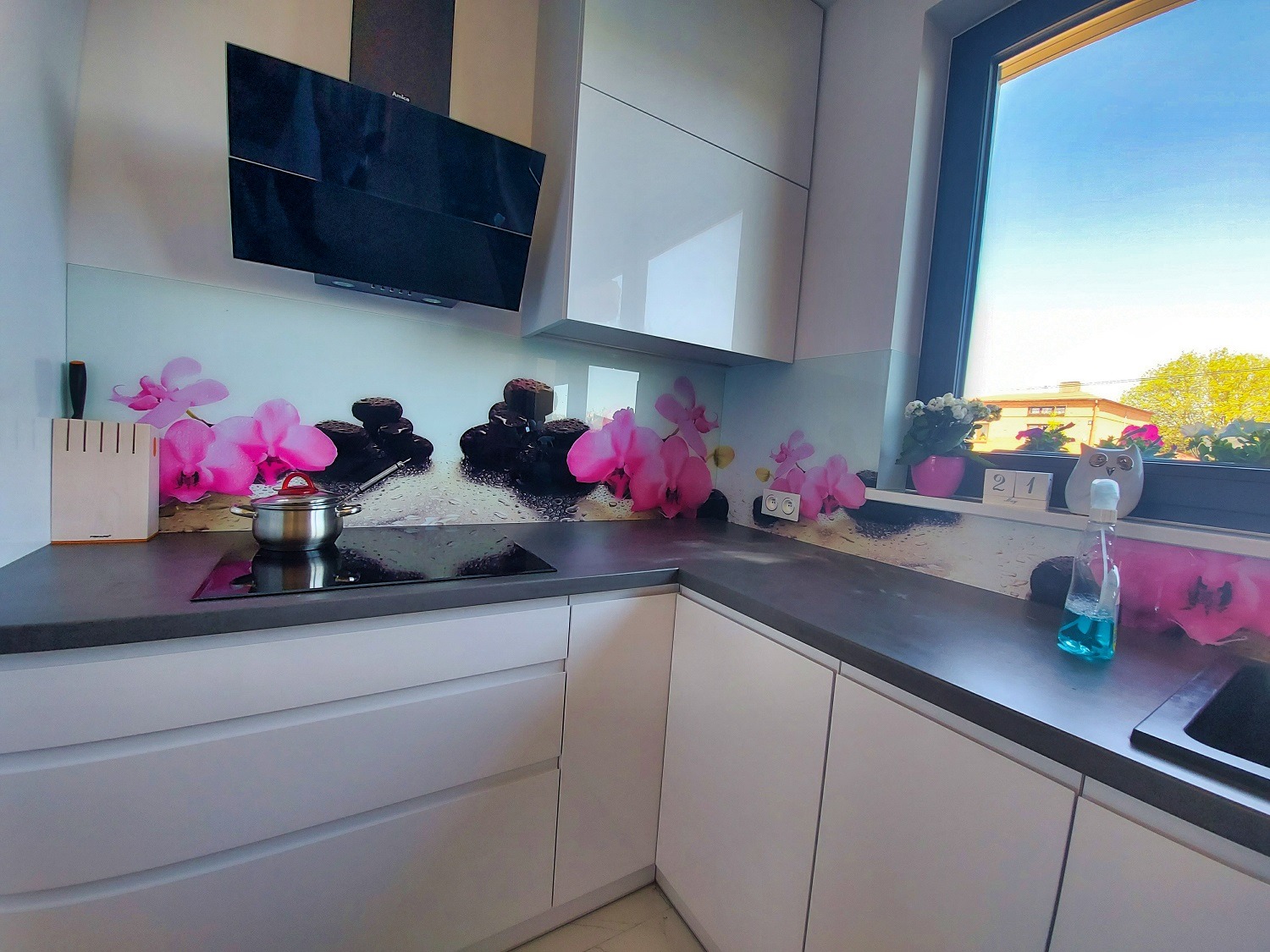 Nowoczesna kuchnia z białymi szafkami i szklanym panelem nad blatem z motywem orchidei i kamieni zen. Na blacie indukcja i garnek. Duże okno z kwiatami.