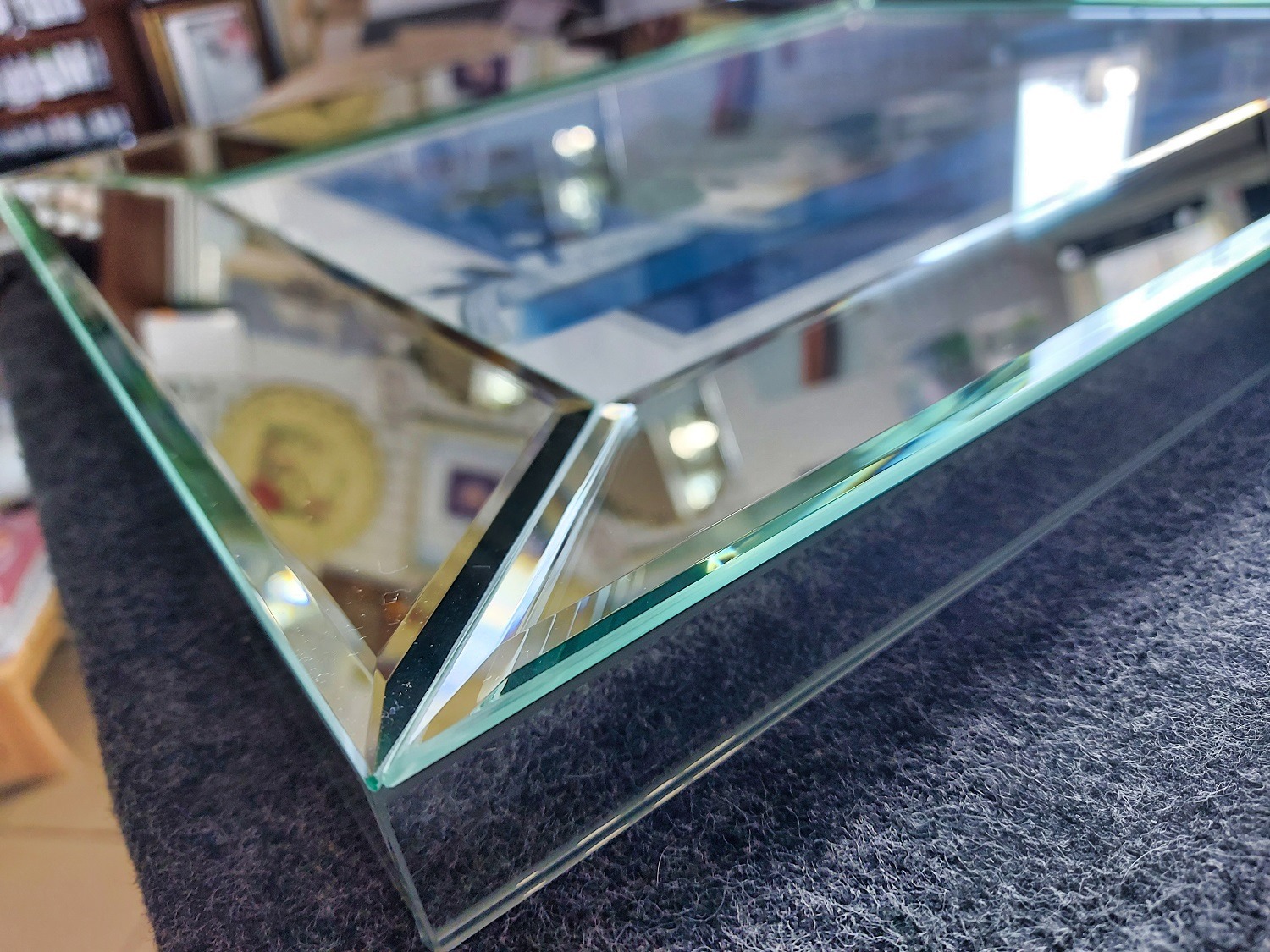 Szklana tafla z fazowanymi krawędziami, leżąca na szarym materiale. Widoczne refleksy światła i otoczenia na powierzchni szkła. Detal wykończenia.