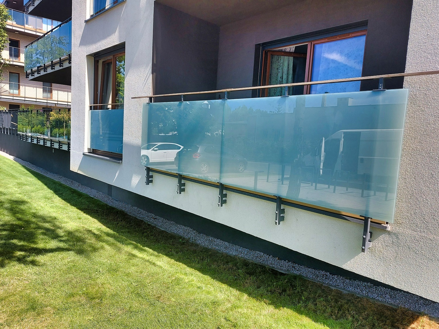 Nowoczesny balkon z szklaną balustradą o błękitnym odcieniu, z widokiem na zieleń trawnika i zaparkowane samochody, mocowany do elewacji budynku w minimalistycznym stylu.