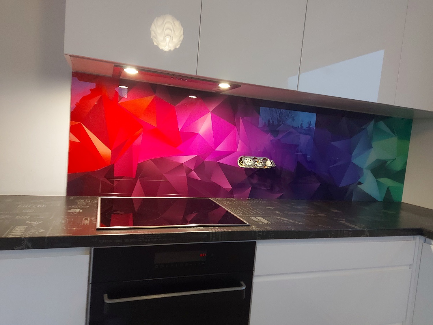 Kolorowy panel szklany nad indukcją, geometryczny wzór w odcieniach czerwieni, fioletu i zieleni. Białe szafki kuchenne i czarny blat z napisami.