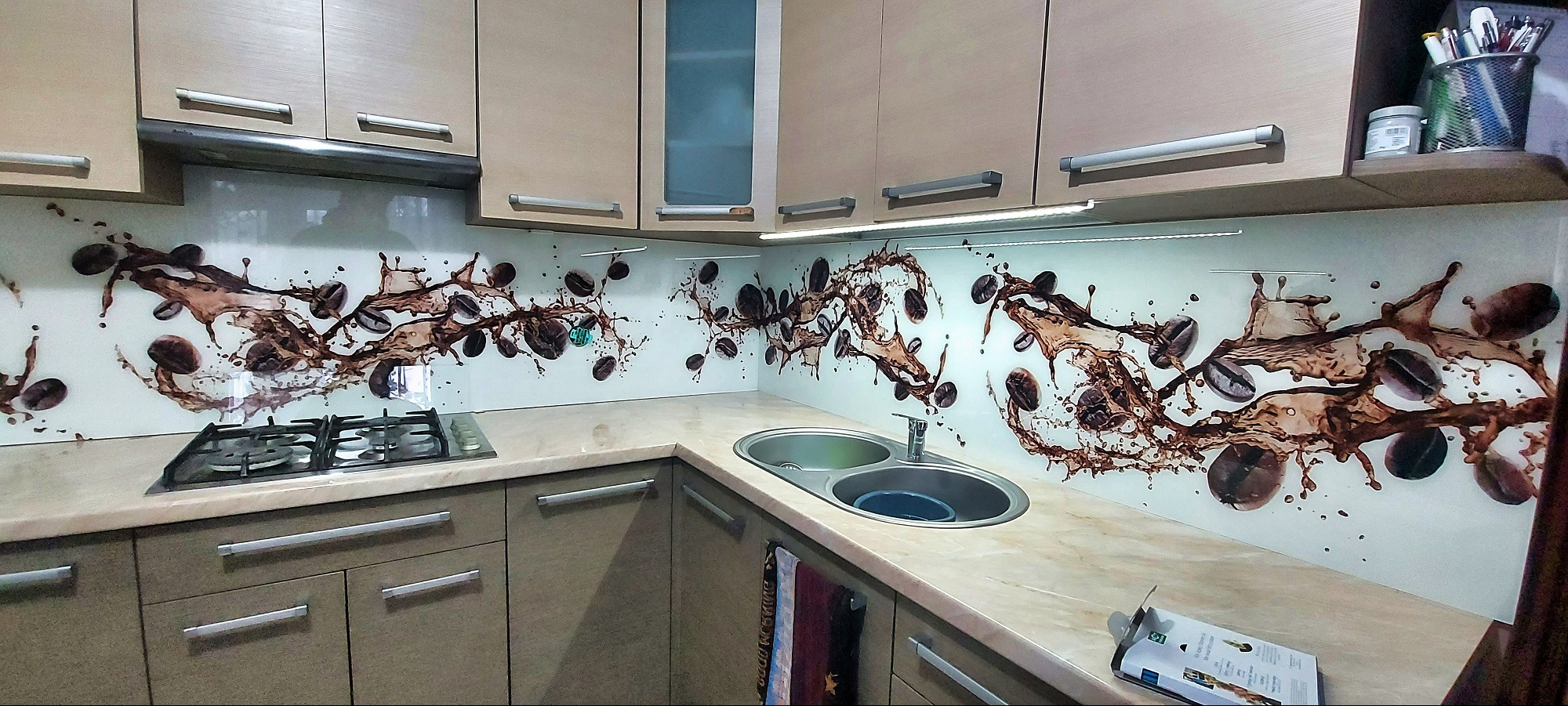 Kuchnia z beżowymi szafkami i szklanym panelem nad blatem z motywem kawy. Nowoczesny wystrój, oświetlenie LED pod szafkami.