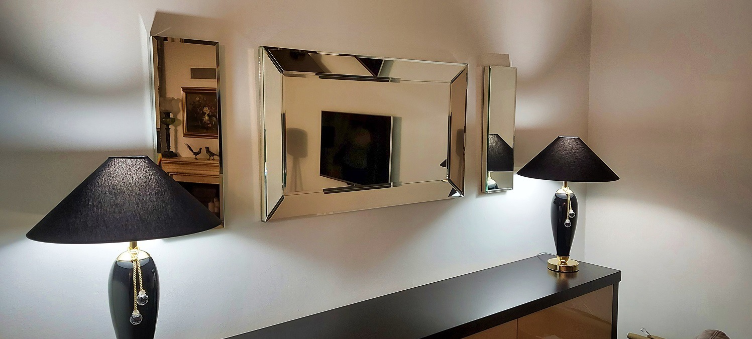 Eleganckie lustra dekoracyjne na ścianie w salonie, odbijające wnętrze. Dwie czarne lampy stołowe z abażurami doświetlają aranżację. Minimalistyczny design.