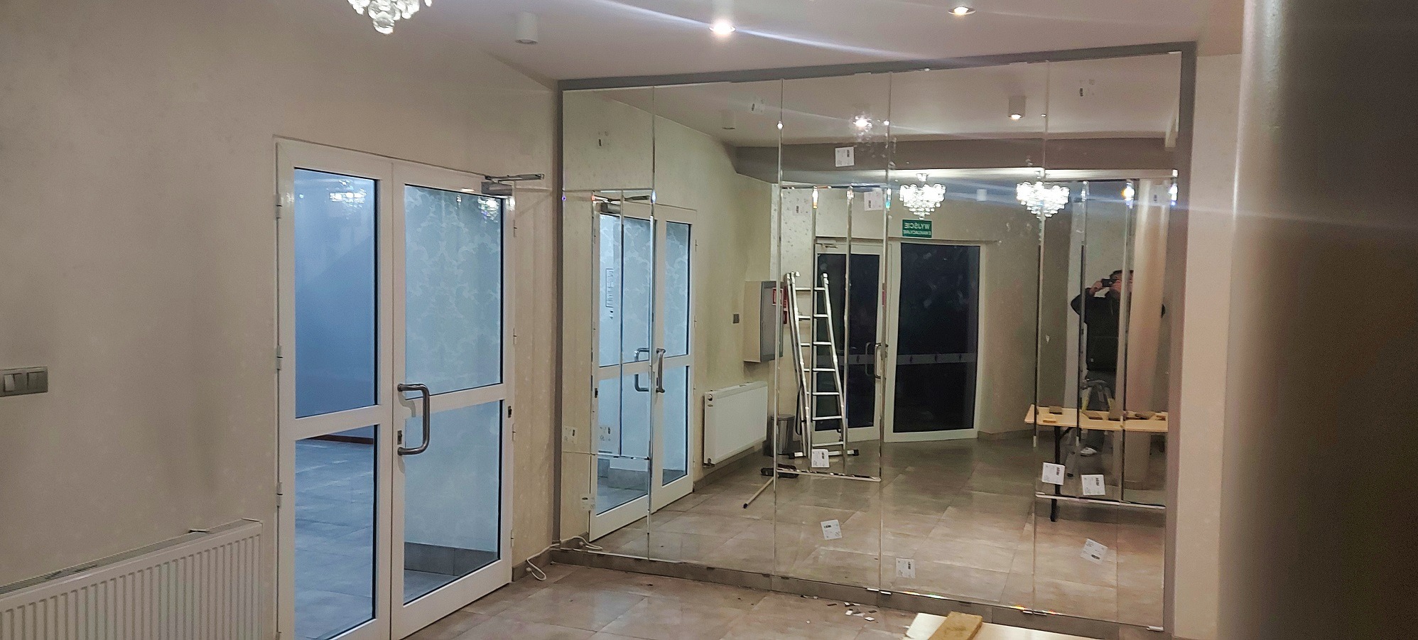 Montaż dużych, lustrzanych paneli ściennych w korytarzu. Widoczne białe drzwi i drabina. Osoba robi zdjęcie w odbiciu lustra. Oświetlenie sufitowe.
