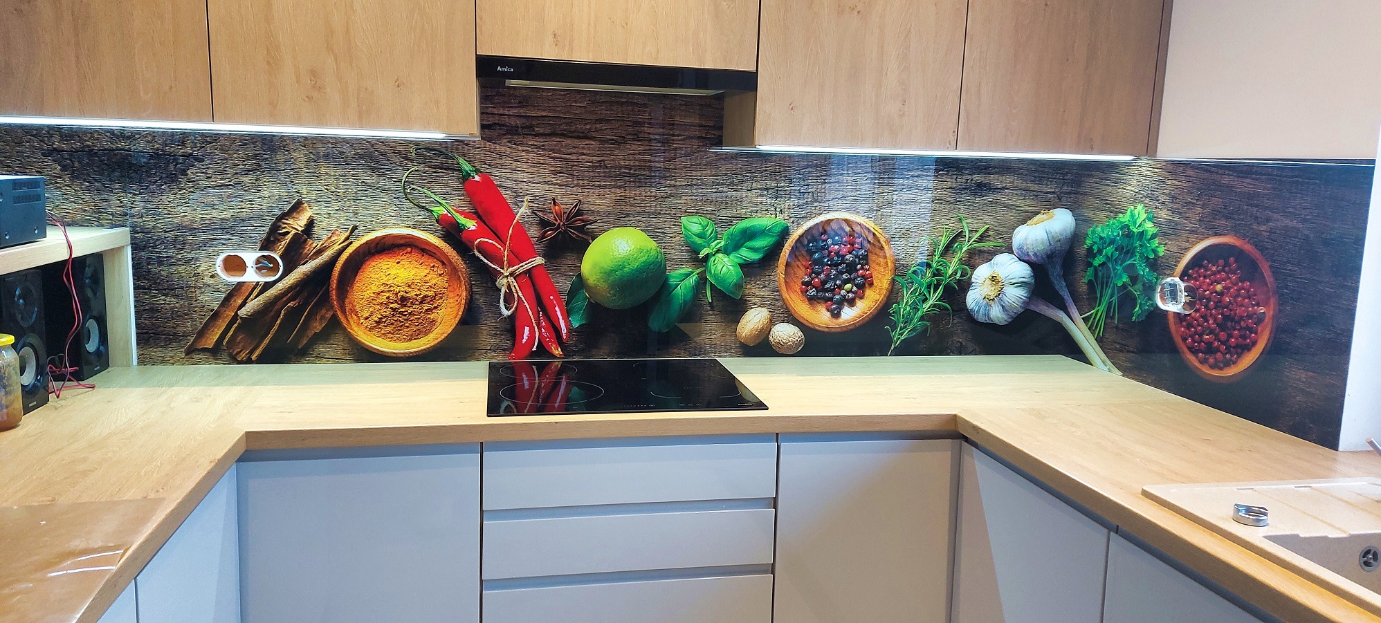 Nowoczesna kuchnia z panelem szklanym nad blatem, z motywem przypraw i ziół. Jasne szafki górne i dolne, drewniany blat. Widoczny fragment instalacji elektrycznej.