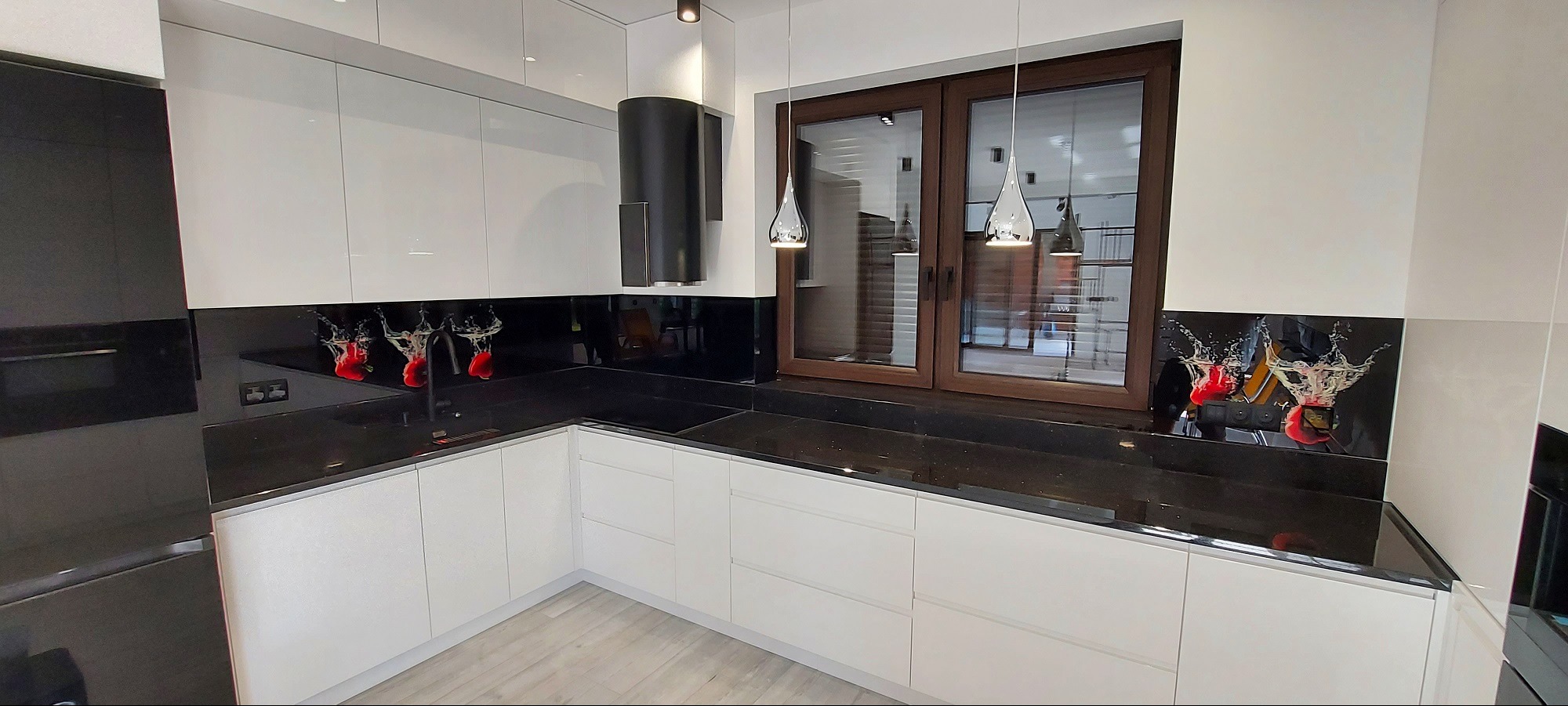 Nowoczesna kuchnia z białymi szafkami, czarnym blatem i panelem szklanym z motywem owoców w wodzie. Widoczne okno w drewnianej ramie i designerskie lampy.