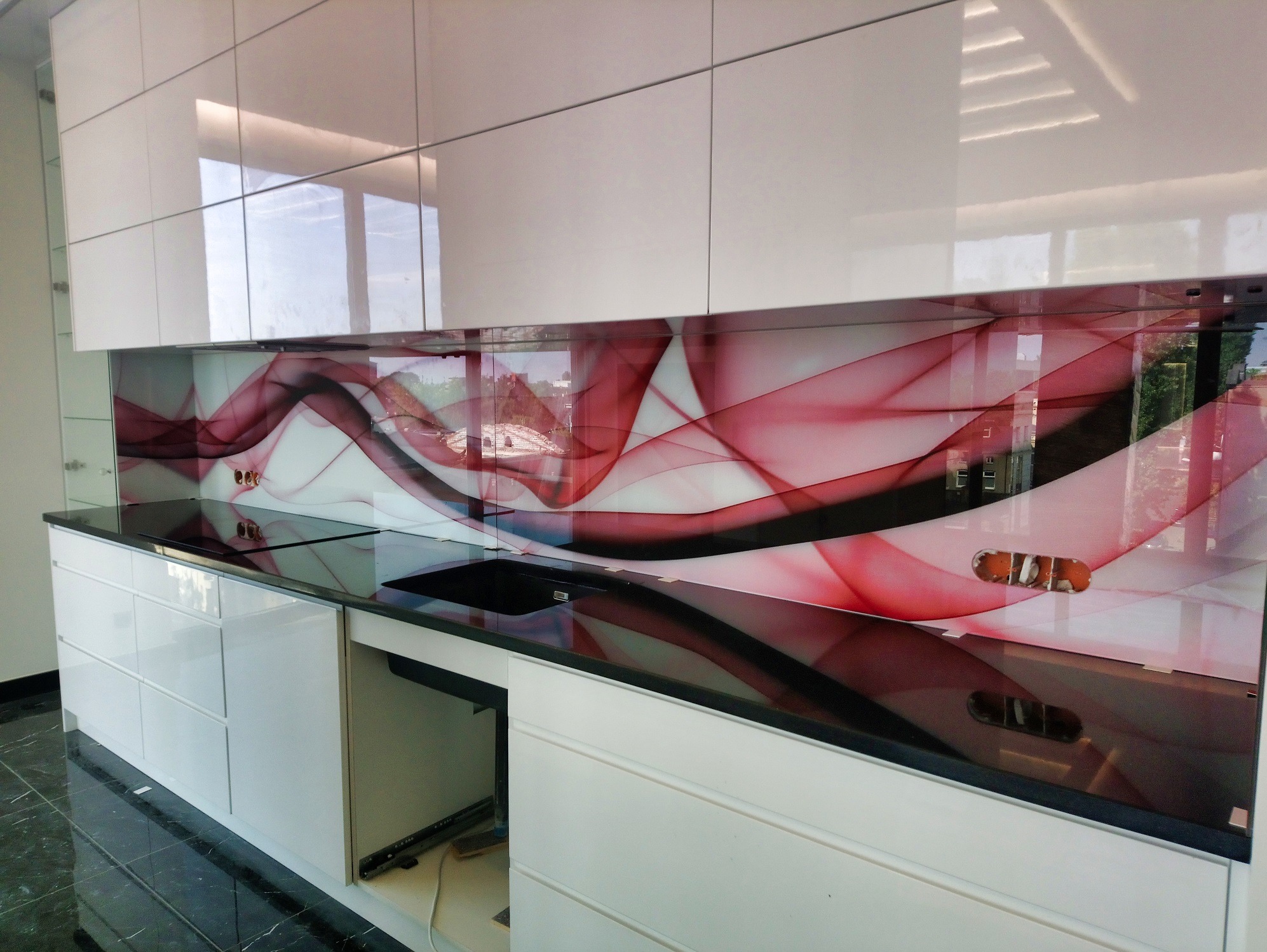 Szklany panel kuchenny z abstrakcyjnym wzorem w odcieniach czerwieni i czerni, zamontowany między białymi szafkami, z widocznymi gniazdkami elektrycznymi.