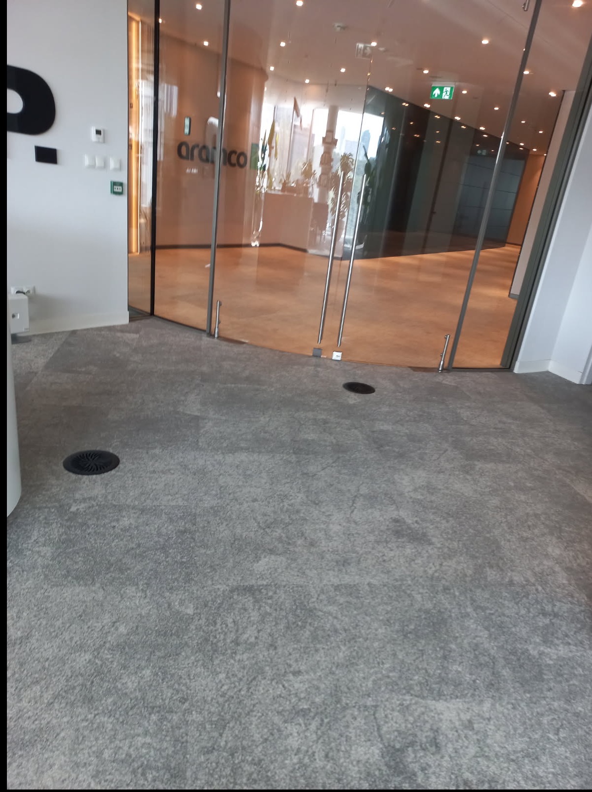 Wnętrze biura z szarą wykładziną i przeszklonym wejściem do pomieszczenia z logo firmy Aramco. Widoczne kratki wentylacyjne w podłodze i jasne oświetlenie.