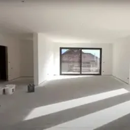 Jasne wnętrze po gładziowaniu ścian i sufitu. Widoczne duże okno przesuwne i dwa mniejsze okna. Podłoga przygotowana do dalszych prac wykończeniowych.