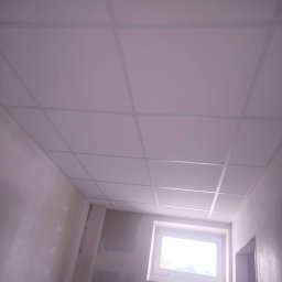 Mario Mariusz Reszka - Biały sufit kasetonowy z widoczną siatką profili, w tle fragment ściany po szpachlowaniu, okno z jasnym światłem dziennym. Ujęcie z dołu.