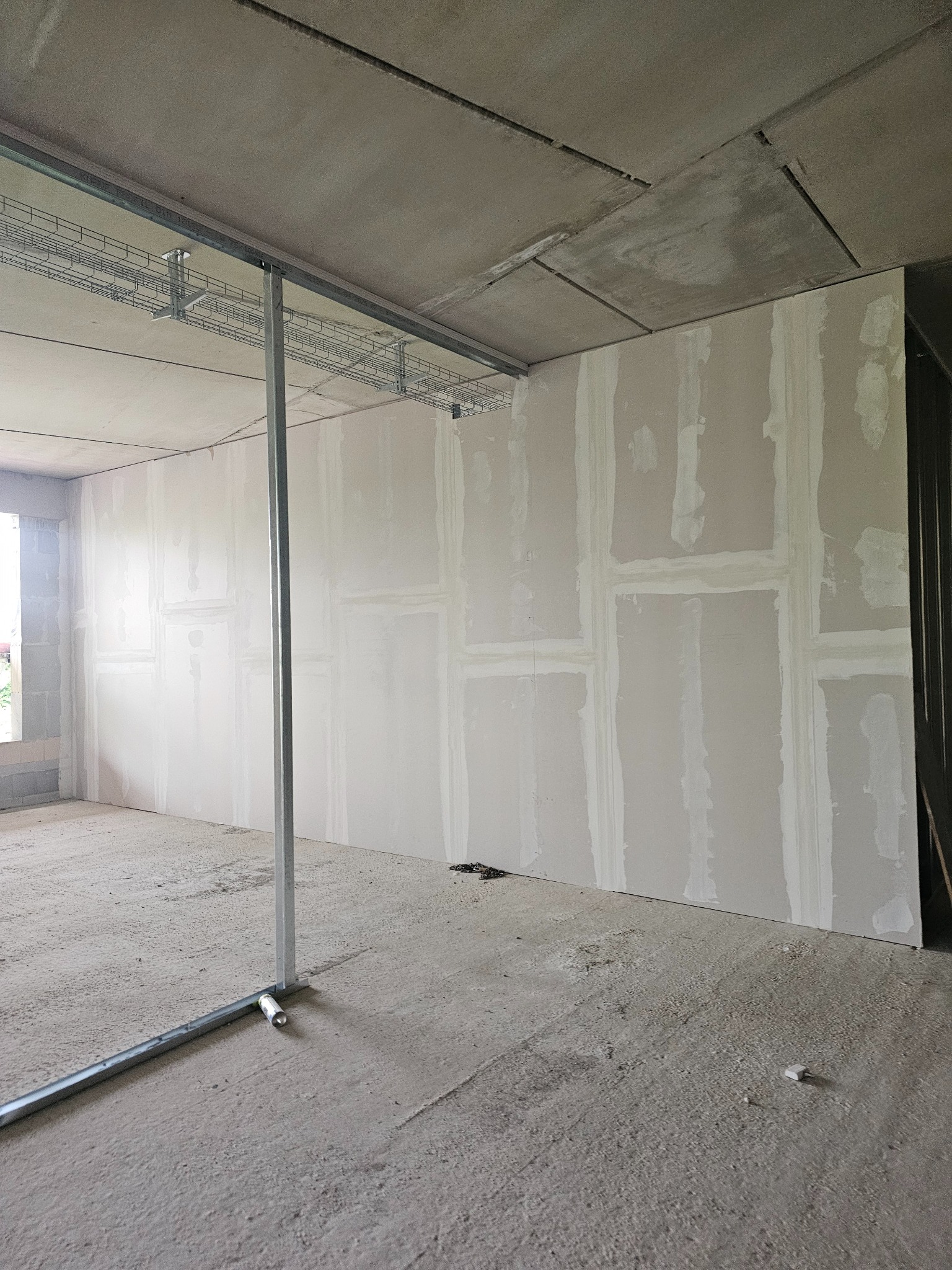 Montaż płyt gipsowo-kartonowych na ścianie z widocznymi łączeniami szpachlowanymi. Metalowa konstrukcja sufitu i podłogi. Surowy stan wykończenia wnętrza.