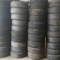 AUTO-KLIMA KLIMATYZACJE SANOK, OPONY UŻYWANE, OPONY BIEŻNIKOWANE I NOWE, PROSTOWANIE ALUFELG SANOK, WYMIANA OPON, WULKANIZACJA SANOK - Używane opony samochodowe ułożone w stosy, widoczne żółte oznaczenia rozmiaru i typu na niektórych oponach, w tle ściana magazynu.