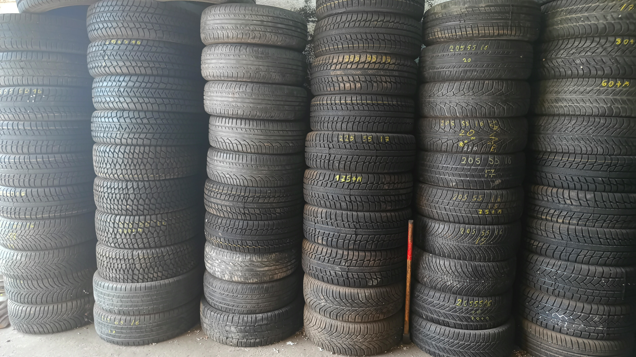 Używane opony samochodowe ułożone w stosy, widoczne żółte oznaczenia rozmiaru i typu na niektórych oponach, w tle ściana magazynu.