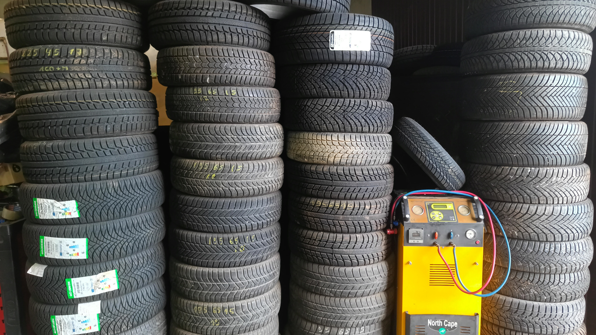 Stosy opon zimowych i letnich z żółtym urządzeniem do obsługi klimatyzacji samochodowej North Cape na pierwszym planie.