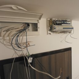 Mikołaj Kryszczuk - Dwie skrzynki elektryczne na białej ścianie. Jedna otwarta z widocznymi kablami, druga zamknięta. Kabel wystaje spod szafki RTV. Ujęcie z dołu.