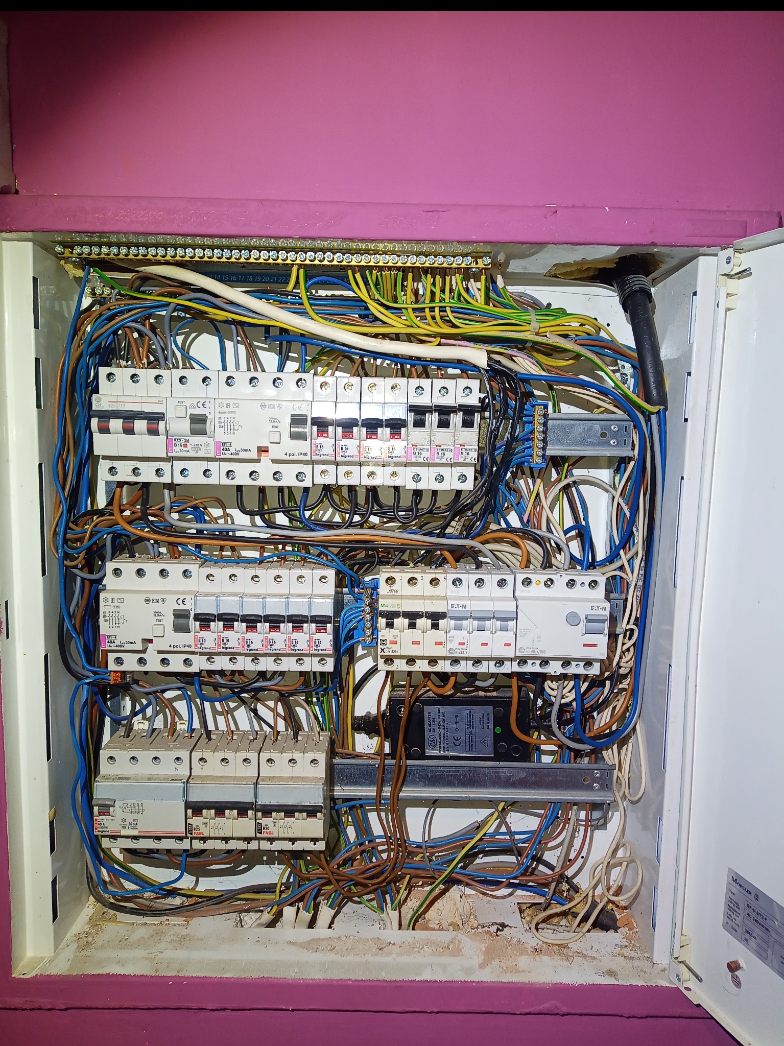 Otwarta skrzynka elektryczna z licznymi bezpiecznikami i przewodami w kolorach: niebieskim, żółtym, brązowym i czarnym. Widoczne elementy montażowe i oznaczenia na urządzeniach.