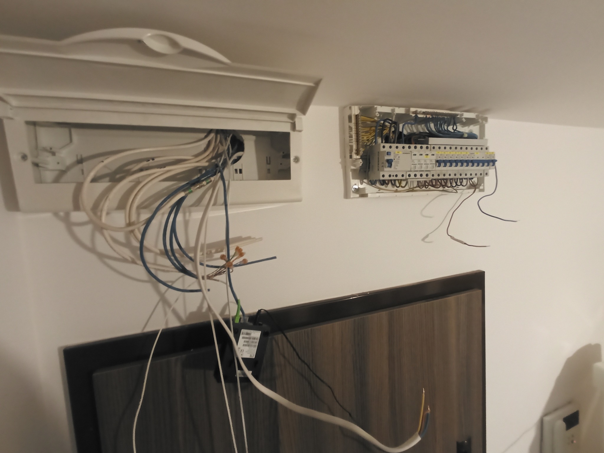 Dwie skrzynki elektryczne na białej ścianie. Jedna otwarta z widocznymi kablami, druga zamknięta. Kabel wystaje spod szafki RTV. Ujęcie z dołu.