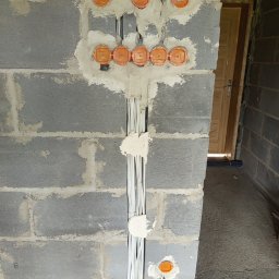 PowerDom - Szara ściana z betonu komórkowego z pomarańczowymi puszkami elektrycznymi i przewodami w bruzdach, zabezpieczonymi gipsem. Widoczny fragment podłogi i drzwi.