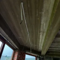 Surowy strop z wyprowadzonymi kablami elektrycznymi. Widoczne okna i ściany z cegły oraz bloczków betonowych. Etap prac instalacyjnych w nowym budynku.