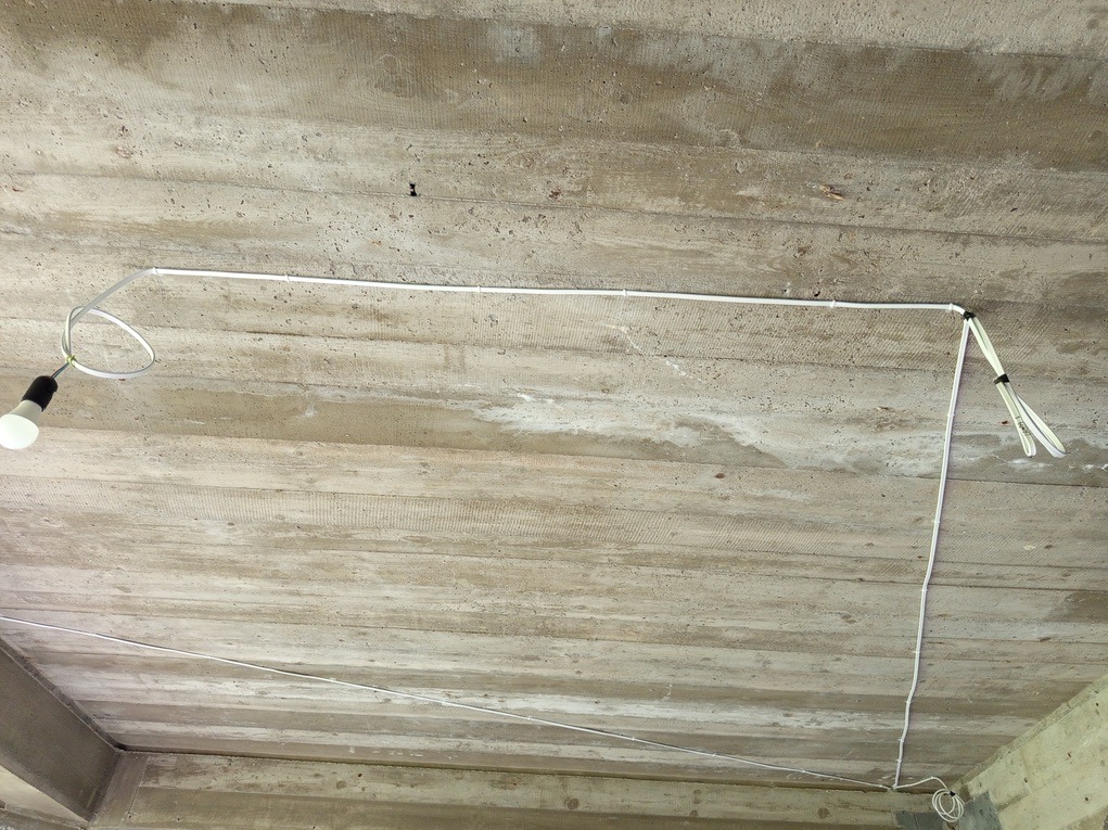 Surowy betonowy strop z prowizorycznym oświetleniem: żarówka na kablu i białe kable ułożone wzdłuż krawędzi stropu. Widoczne łączenia kabli.