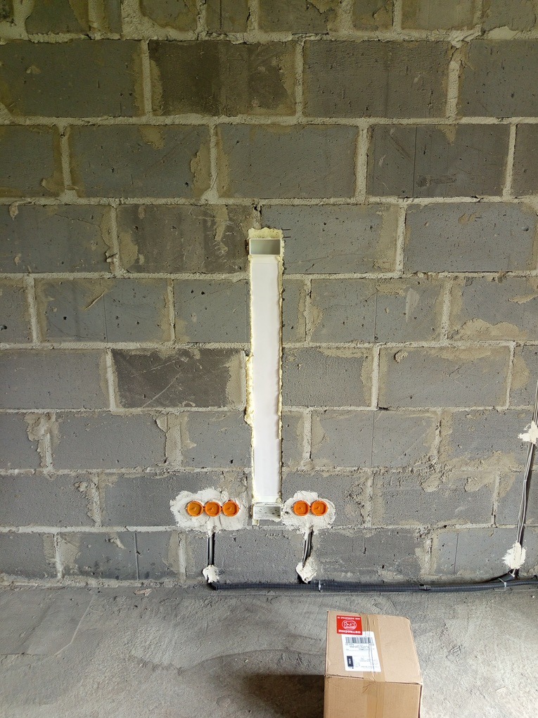 Wykute bruzdy w szarej ścianie z betonu komórkowego, w nich rurki instalacyjne i puszki elektryczne zalane gipsem. Widoczny przewód i karton na podłodze.