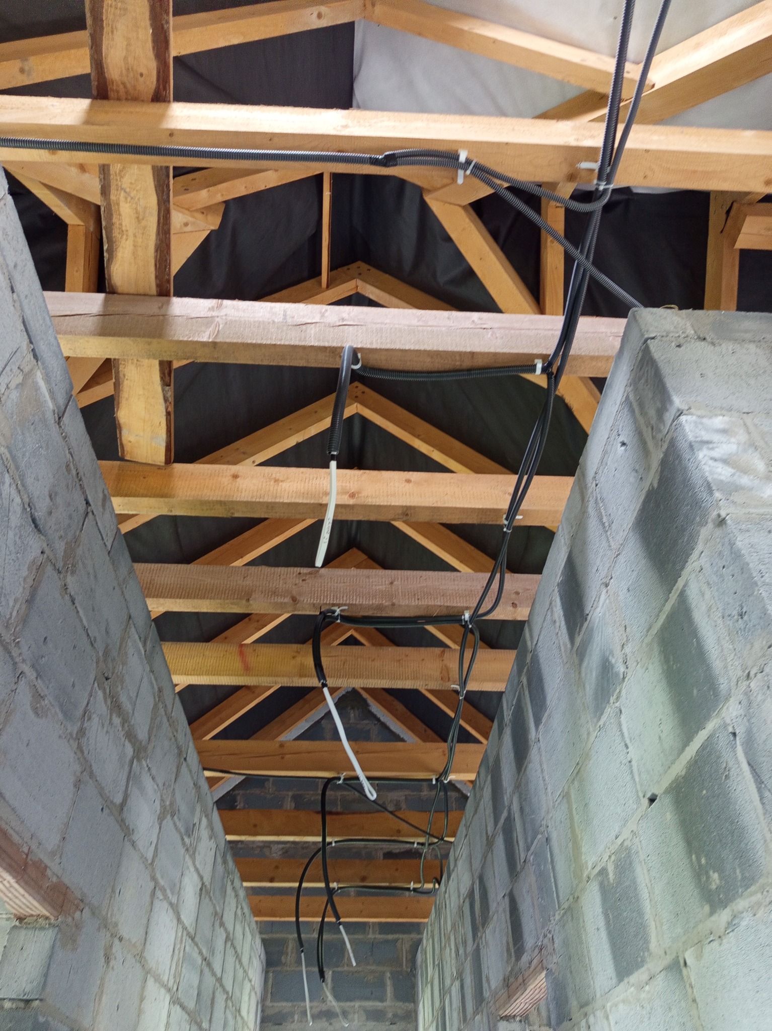 Wnętrze w trakcie budowy: surowa konstrukcja dachu z drewnianymi belkami i czarną folią, widoczne przewody elektryczne zamocowane do belek i murowane ściany z bloczków.
