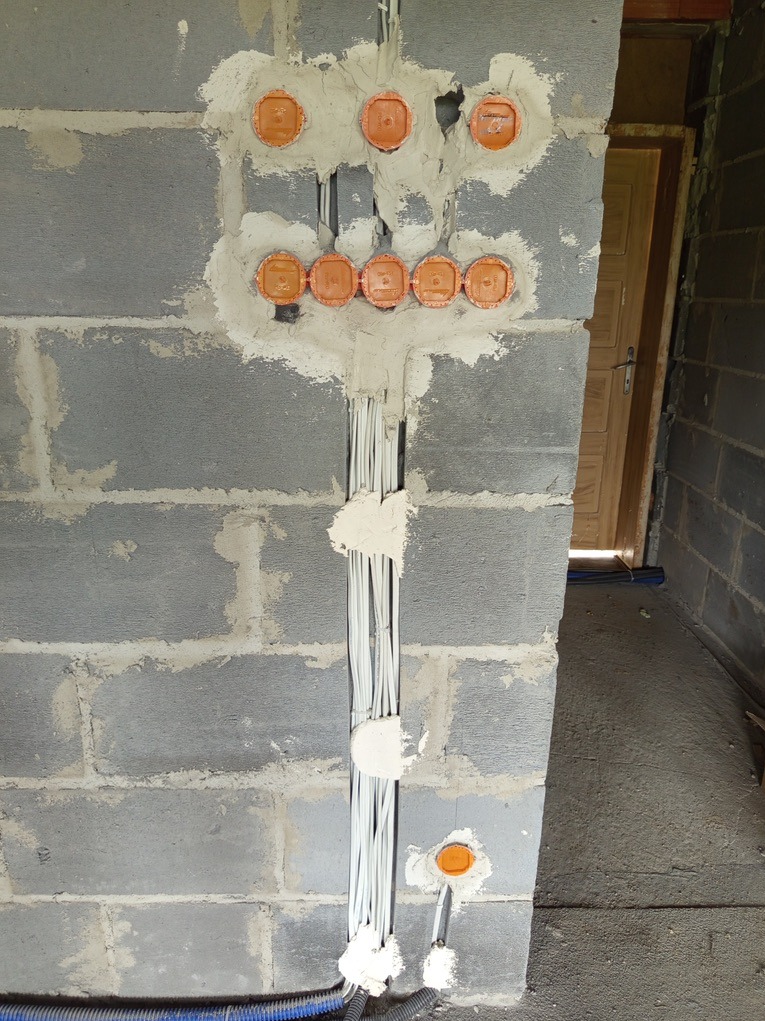 Szara ściana z betonu komórkowego z pomarańczowymi puszkami elektrycznymi i przewodami w bruzdach, zabezpieczonymi gipsem. Widoczny fragment podłogi i drzwi.