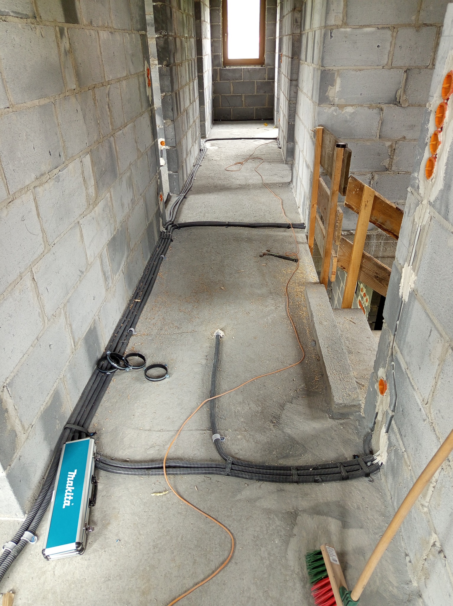 Wnętrze w trakcie prac: szare ściany z betonu komórkowego, rury instalacji elektrycznej w korytach, narzędzia i kufer Makita na podłodze. Pomarańczowe puszki elektryczne.