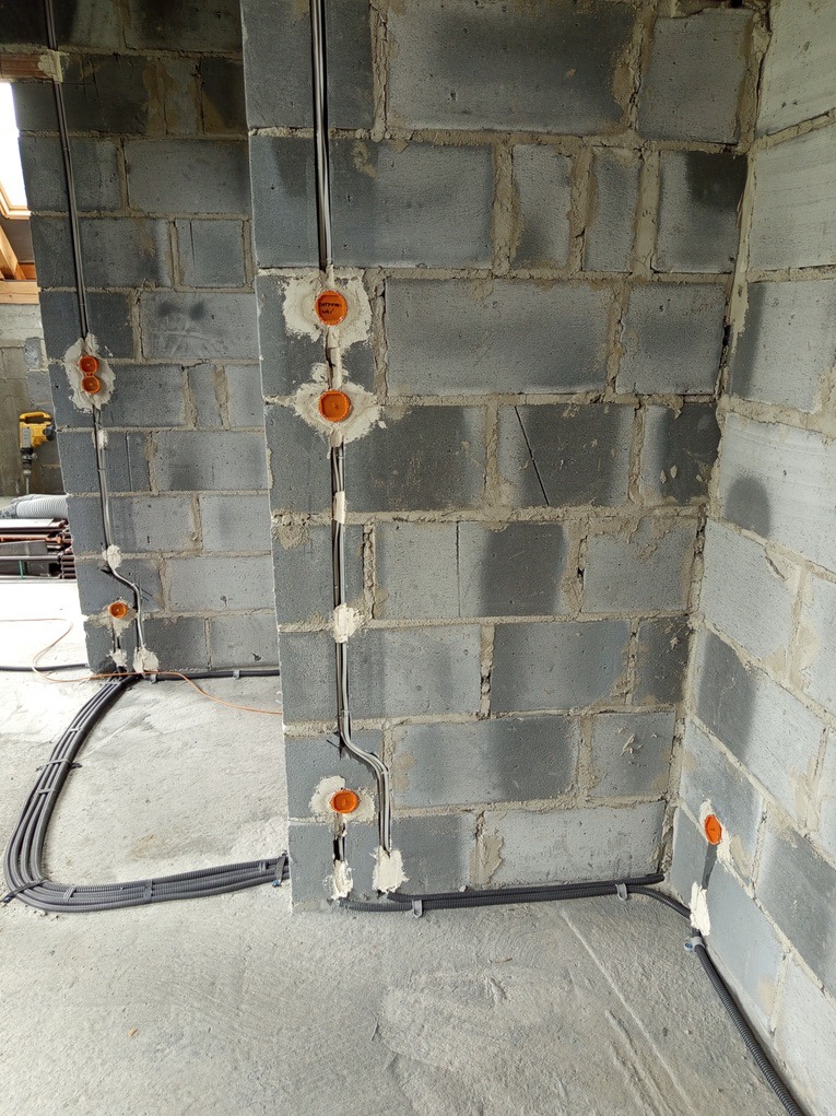 Szara ściana z betonu komórkowego z zamontowanymi puszkami elektrycznymi i poprowadzonymi przewodami w peszlach, zabezpieczonymi gipsem.