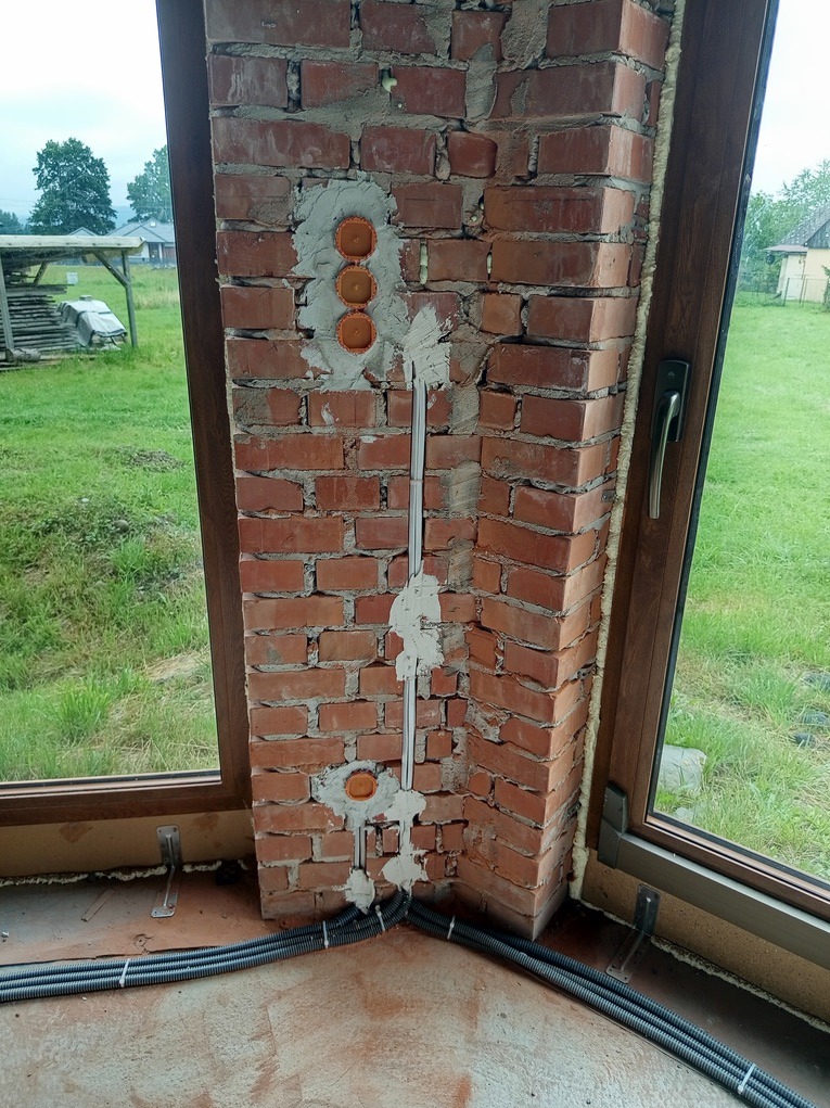 Ceglana ściana z zamontowanymi puszkami elektrycznymi i przewodami w szarych rurkach. W tle widoczne okno z widokiem na zewnątrz.
