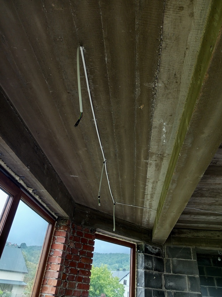Surowy strop z wyprowadzonymi kablami elektrycznymi. Widoczne okna i ściany z cegły oraz bloczków betonowych. Etap prac instalacyjnych w nowym budynku.