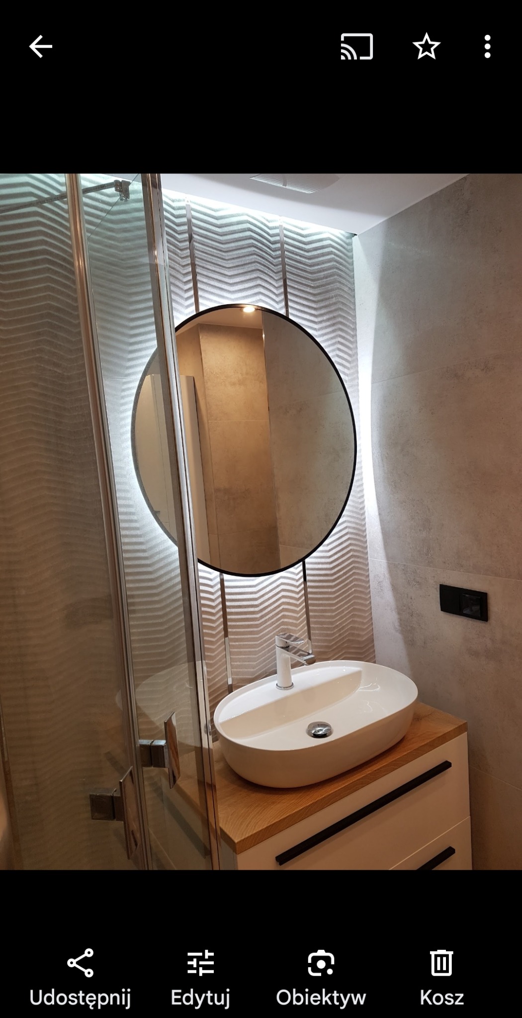 Stylowa łazienka z okrągłym lustrem LED, umywalką nablatową na drewnianym blacie i szklaną kabiną prysznicową. Dekoracyjne płytki 3D na ścianie.