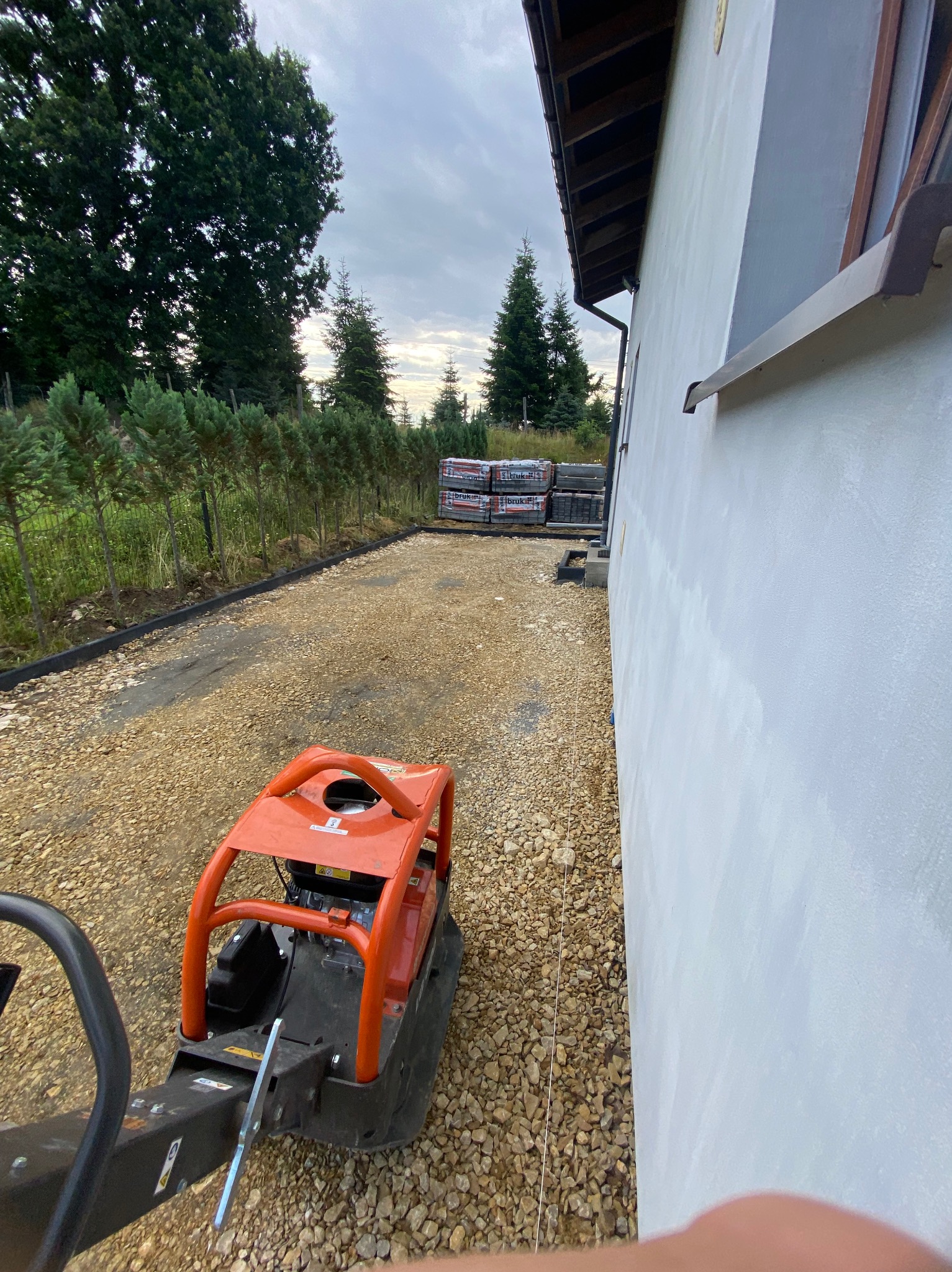 Prace brukarskie: zagęszczarka wibruje tłuczeń na podjeździe. W tle kostka brukowa i rząd tuj. Widok na dom w pochmurny dzień.